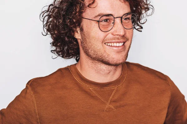 Closeup horizontal portrait of handsome smiling young male with curly hair, wears brown pullover and round trendy spectacles, looking to the camera. Business man posing on white studio wall. Copyspace — Stock Photo, Image