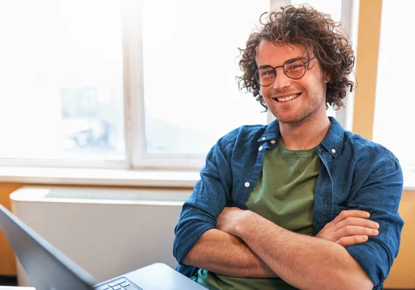 Binnenshuis shot van gelukkig jongeman glimlachend en werken op de computer in de moderne werkplek tegen het raam. Jonge ondernemer werken bij het opstarten, stak zijn handen en kijkt naar de camera. — Stockfoto