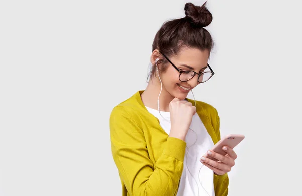 Indoor portrait of brunette smiling young woman using cellphone, messaging to her friend. Happy female chatting with her boyfriend, looking at screen of smart phone and earphones. — Stock Photo, Image