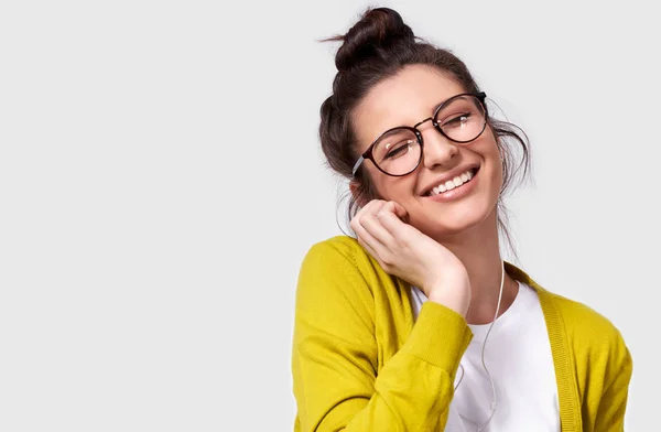 Studio horisontella närbild porträtt av uppriktig leende kvinna i glasögon röra hörlurar. Inomhus skott av kaukasiska ung kvinna med glansig hy bär gula blusen och vit t-shirt. Kopiera utrymme — Stockfoto