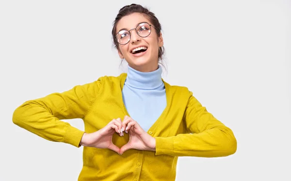 Imagen de una joven europea sonriente haciendo forma de corazón con los dedos. Hermosa sonrisa femenina ampliamente en suéter azul y amarillo que muestra el gesto del corazón con dos manos, signo de amor, sobre fondo blanco . —  Fotos de Stock