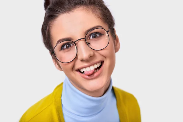 Closeup studio portrait of optimistic funny woman with bun hairstyle showing her tongue and wearing round trendy spectacles while posing on white studio wall. Positive smiling female making grimace — Stock Photo, Image