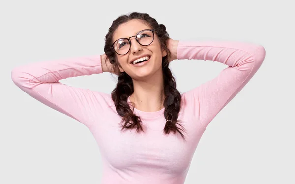 Positiv kaukasisk kvinna student håller händerna på huvudet, brett leende, bär Rosa casual kläder och runda glasögon, isolerade över vit bakgrund. Glad ung kvinna med flätor frisyr på vit — Stockfoto