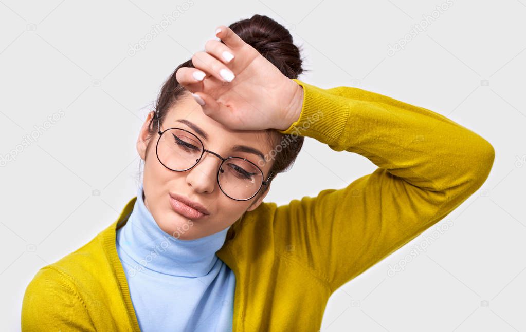 Tired pretty brunette woman in bun and wearing casual outfit with round trendy eyeglasses, keeps hand on forehead, while standing over white wall. People and amotion concept
