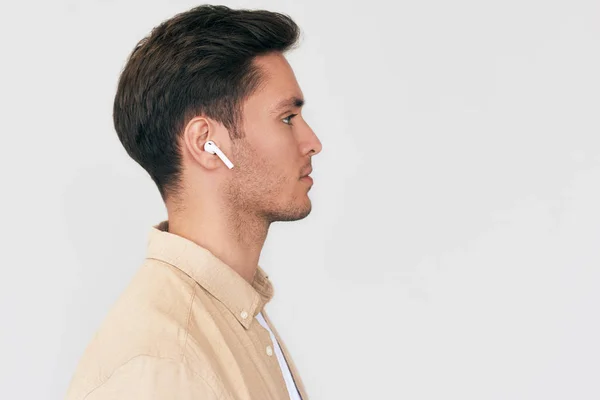 Side view portrait of serious young handsome man posing with wireless earphones on white studio background. Caucasian businessman using wireless headphone. — Stock Photo, Image
