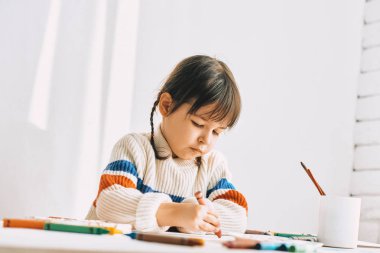 Petrol kalemler ile resim, evde beyaz masa başında oturan sevimli küçük kız ressam portresi. Çok okul öncesi çocuk ile renkli kalemler çizer. Kişi, çocukluk ve eğitim kavramı