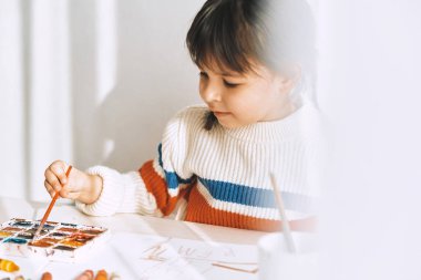 Sevimli küçük kız boyama ve bir fırça ve suluboya ile evde beyaz masanın üzerinde çizim. Çok okul öncesi çocuk gülümseyen anaokulunda çizer. Kişi, çocukluk ve eğitim kavramı