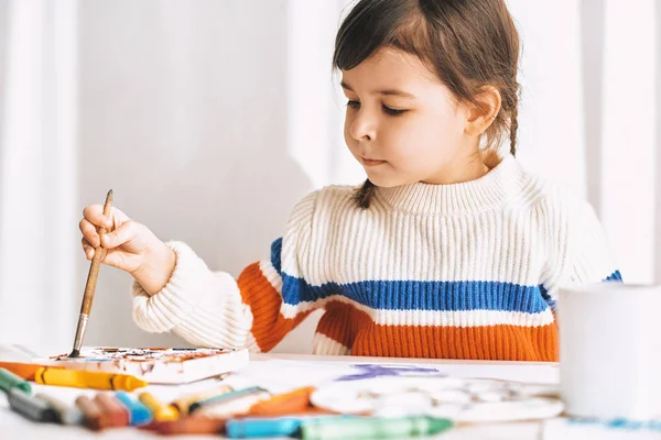 Художественная маленькая девочка живопись и рисунок с карандашами и акварелью на белом столе дома. Улыбающийся ребенок в детском саду рисует кисточкой. Человек, детство и воспитание — стоковое фото