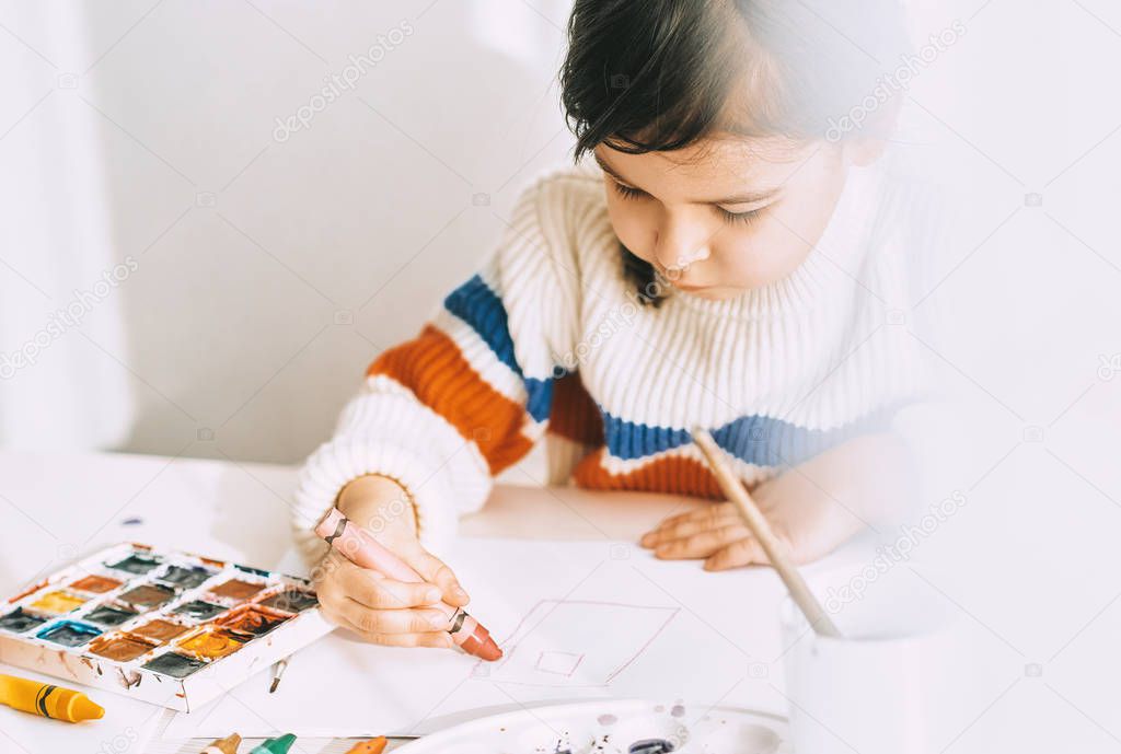 Indoor image of artistic little girl painting with oil pencils, sitting at white desk at home. Pretty smiling preschool kid drawing with pencils. People, childhood and education concept