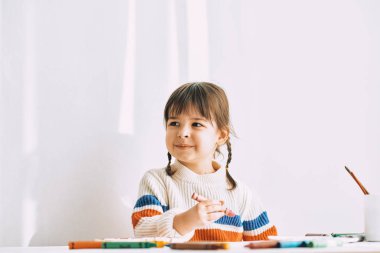 Yatay görüntü mutlu sevimli küçük kız evde beyaz masa başında oturan petrol kalemler ile boyar. Çok okul öncesi çocuk gülümseyen kalemler ile çizer. Uzay kopyalayın. Kişi, çocukluk ve eğitim kavramı