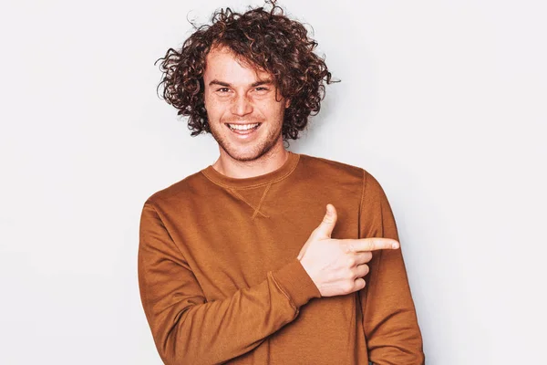 Hombre joven feliz con el pelo rizado lleva jersey indica un lado con el dedo delantero, sonriendo y mirando a la cámara, demuestra algo en espacio de copia en blanco para la promoción. Gente, concepto de emoción — Foto de Stock