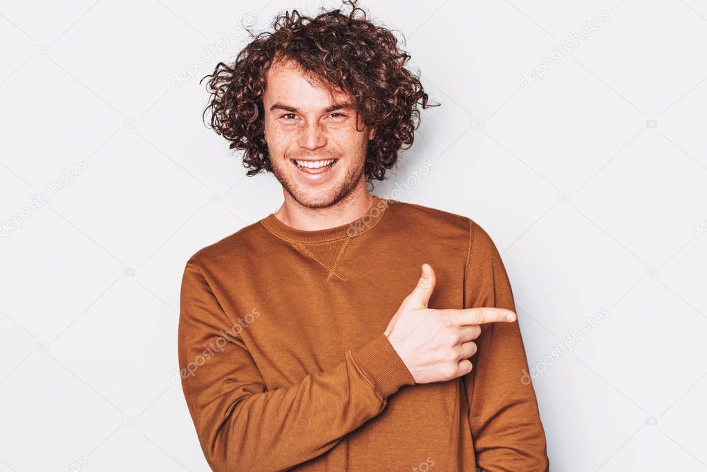Happy young male with curly hair wears pullover indicates a side with fore finger, smiling and looking at the camera, demonstrates something at blank copy space for promotion. People, emotion concept
