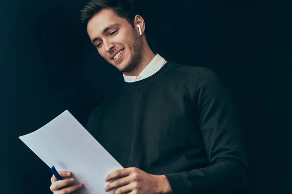 Šťastný mladý pohledný muž s úsměvem a čtení informací z documanet a rozhovor z bezdrátové sluchátka. Podnikatel, používající bezdrátová sluchátka na konferenci dělat poznámky na papír. — Stock fotografie