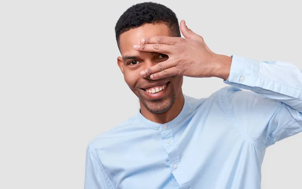 アフリカ系アメリカ人のハンサムな若い男は手のひらで顔を隠すと、彼の目を見る。青い t シャツを着て、ポーズを白い壁にカメラを見て手で顔を覆う若い男性のスタジオ ポートレート — ストック写真