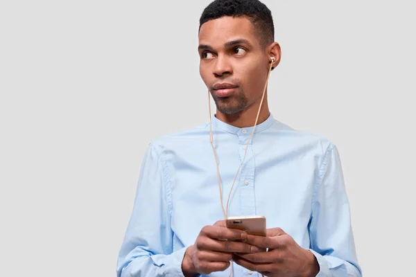 アフリカ系アメリカ人の若い男性の肖像画は、現代のスマート フォン、携帯電話でブログを保持しています。ハンサムな男性は、携帯電話経由でイヤホンで音楽を聴きます。人と技術の概念 — ストック写真