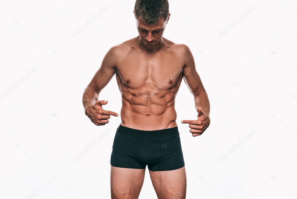 Handsome male model in black underwear showing his sexy body, standing on a white background. Shirtless bodybuilder man with sexy abdomen and biceps posing over white wall. Health and people concept