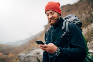Young bearded man smiling and sending messages for his family from his cellphone, during hiking in mountains. Traveler bearded man in red hat using mobile phone application. Travel and lifestyle clipart