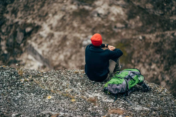 Colpo in testa di giovane maschio rilassarsi in montagna dopo l'escursionismo. Vista posteriore di viaggiatore uomo in cappello rosso, seduto da solo hanno una pausa dopo l'alpinismo. Viaggio e stile di vita — Foto Stock