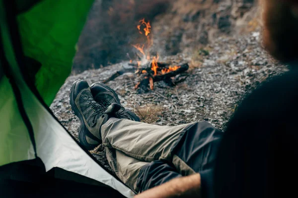 Ausgeschnittenes Bild vom Zelteingang eines jungen Reisenden, der sich in den Bergen in der Nähe des Lagerfeuers entspannte. Außenaufnahme eines Männchens, das im Zelt sitzt und nach dem Wandern zum Feuer blickt. Reise — Stockfoto