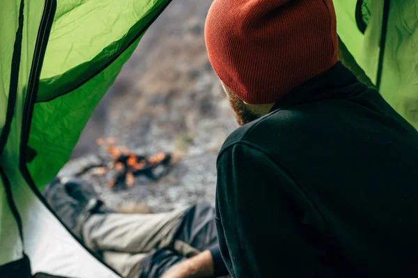 Bild von einem Blick vom Zelteingang eines jungen Reisenden, der sich in den Bergen in der Nähe des Lagerfeuers entspannte. Aufnahme von hinten, wie ein männlicher Wanderer im Zelt sitzt und nach dem Wandern zum Feuer blickt. Reise — Stockfoto