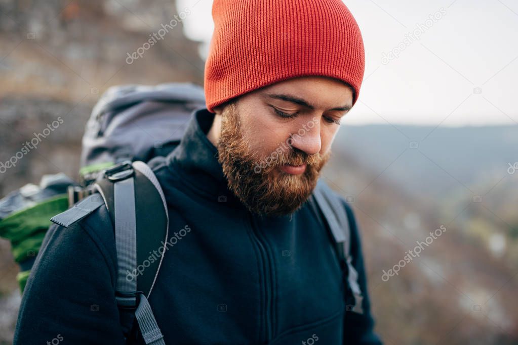 Closeup outdoors portrait of young handsome hiker male hiking in mountains wearing red hat. Traveler bearded man trekking and mountaineering. Travel, people and healthy lifestyle concept