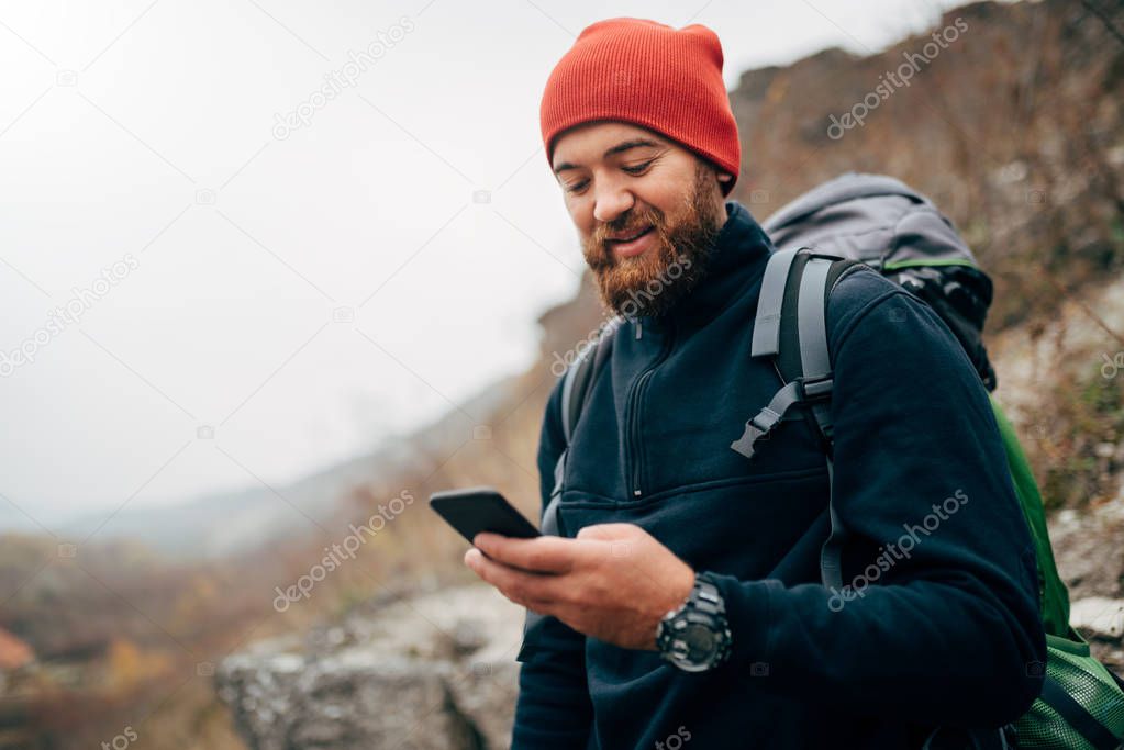 Young bearded man smiling and sending messages for his family from his cellphone, during hiking in mountains. Traveler bearded man in red hat using mobile phone application. Travel and lifestyle