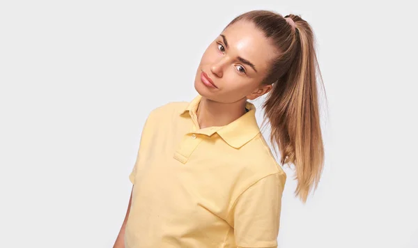 Beautiful blonde young woman with a pony tail looking at camera with cute shy smile, feeling a bit shy or uncomfortable. Pretty female wearing yellow t-shirt on white studio wall. People emotions — Stock Photo, Image