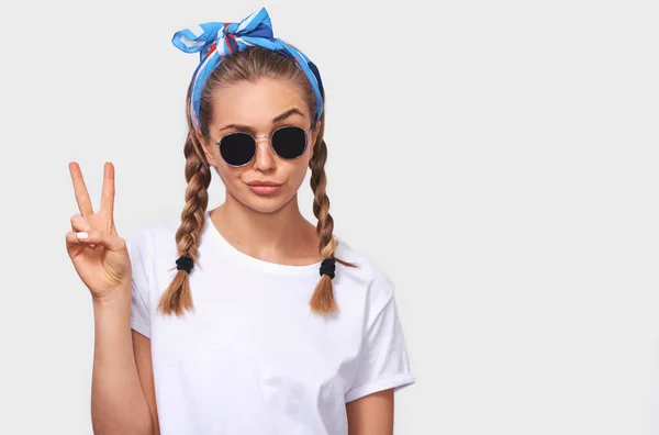 Studioporträt einer fröhlichen blonden jungen Frau mit modischer Sonnenbrille, weißem T-Shirt und blauem Stirnband, die ein Entengesicht macht und ein Friedenszeichen zeigt. Studentin wird mit Zöpfchen-Frisur verrückt — Stockfoto