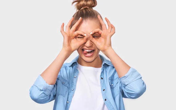 Cerrar imagen horizontal de la joven alegre con el peinado de moño rubio, mostrando gestos Ok con ambas manos, fingiendo llevar gafas ot binocular. Bastante hembra sonriendo y mostrando la lengua . —  Fotos de Stock