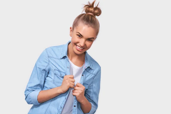 Snygga glada kvinnlig student med bulle frisyr leende positivt och att vara på bra humör när du står över vita studio bakgrund. Ung kvinna som bär blå jeansskjorta brett leende — Stockfoto