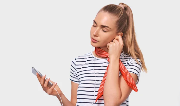 Pretty European young woman with ponytail hairstyle, wearing striped t-shirt, stylish red scarf on neck, relaxing with closed eyes listening to her favourite songs via white earphones using smartphone — Stock Photo, Image