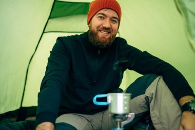 Mutlu genç adam explorer dağlarda sıcak içecek içme portresi. Gezgin, kamp çadır yakın oturan kırmızı şapkalı adam yürüyüş sonra bir fincan çay hazırlama sakallı. Seyahat, yaşam biçimi