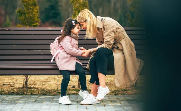 Image horizontale franche de la belle mère et son enfant fille souriant, assis sur un banc à l'extérieur. Aimant jeune femme joyeuse et sa fille heureuse passer du temps ensemble dans le parc de la ville. Fête des mères — Photo