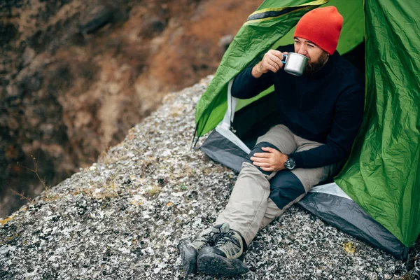 Imagem horizontal ao ar livre do jovem homem empanado bebendo bebida quente nas montanhas. Homem viajante de chapéu vermelho, sentado perto da tenda de acampamento, segurando nas mãos uma caneca de chá depois de caminhar. Viagens, estilo de vida . — Fotografia de Stock