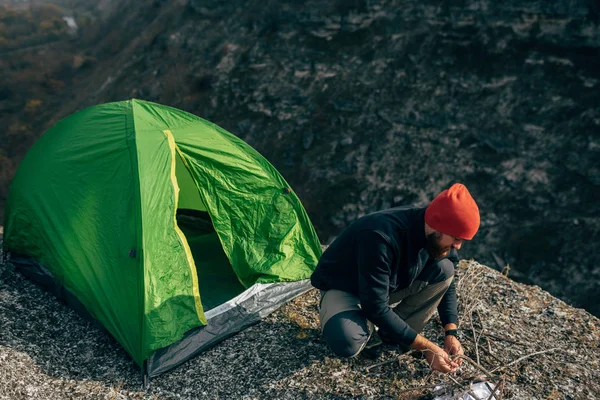 Im Freien bereiten junge Wanderer Brunches für Lagerfeuer in den Bergen vor. Ein bärtiger Mann, der in der Nähe eines Zeltes sitzt und nach dem Wandern ein Lagerfeuer macht. Reisen, Lebensstil — Stockfoto