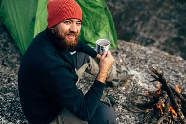 Außenaufnahme von jungen panierten Männern, die in den Bergen Heißgetränke trinken. Reisender mit rotem Hut, der in der Nähe von Zelten und Lagerfeuer sitzt und nach dem Wandern einen Becher Tee in der Hand hält. Reise — Stockfoto