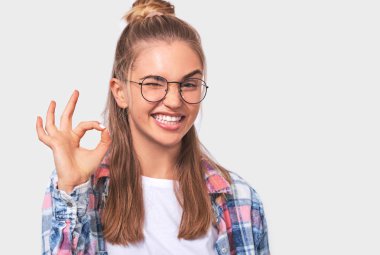 Neşeli mutlu genç kadın giymiş beyaz gömlek, ekose gömlek ve yuvarlak şeffaf gözlük, geniş, gülümseyen iyi jest gösterir, birinin fikri sever, beyaz stüdyo duvar üzerinde pozlar.