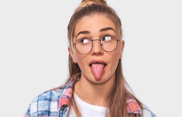 Close up indoors shot of pretty young blonde woman dressed casual outfit and eyewear, making funny faces at camera, sticking out her tongue. Positive female looks aside, shows tongue, making grimace. — Stock Photo, Image