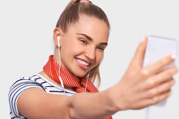 Ritratto ravvicinato di bella giovane donna europea in t-shirt a righe e sciarpa rossa sul collo, scattando autoritratto sopra la parete bianca dello studio. Felice femmina sorridente e prendendo un selfie sul suo smartphone . — Foto Stock
