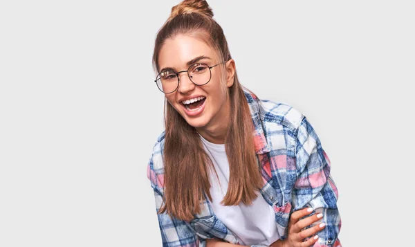 Feliz hermosa mujer joven positiva vestida con traje casual, con gafas redondas transparentes con una agradable sonrisa amplia, mirando a la cámara y posando sobre la pared del estudio blanco. Personas emociones — Foto de Stock