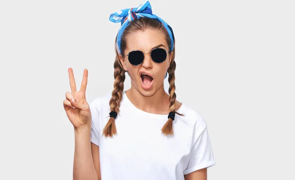 Horizontal studio portrait of cheerful blonde young woman wearing trendy sunglasses, white t-shirt and blue headband, showing peace sign. Student girl going funny with braids hairstyle. People emotion Royalty Free Stock Photos