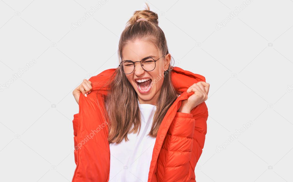 Cheerful blonde young woman screaming with wide open mouth, dressed in white t-shirt and red jacket, wearing round eyewear, isolated over white background. People emotions