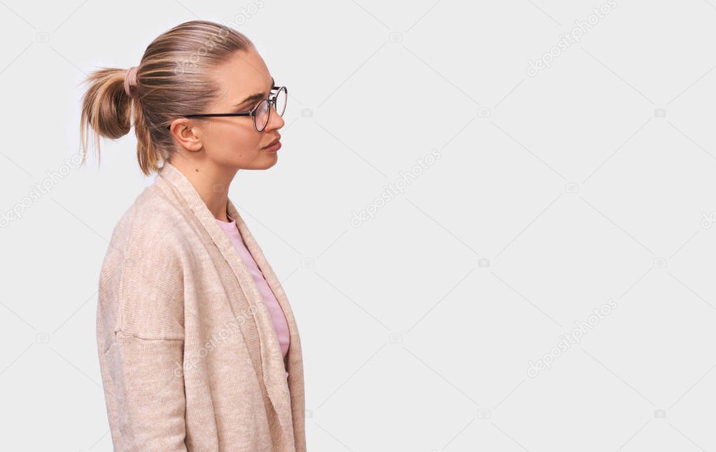 Side view indoor portrait of blonde young woman in eyewear, looking away to the blank copy space, isolated over white wall. Caucasian female with hair bun wearing warm blouse having serious expression