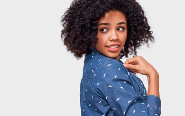 Stüdyo yatay bir yana, beyaz arka plan üzerinde duran bakarak mavi gömlek giymiş güzel mutlu Afrikalı-Amerikalı genç kadın kıvırcık saçlı portresi kapatın. İnsanlar kavramı