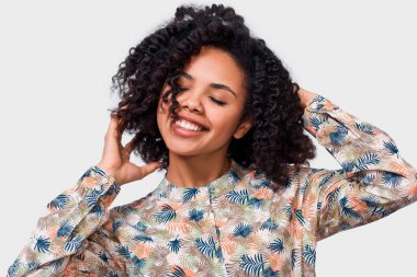 Pleased dark-skinned young woman dressed in floral shirt, holding hands on head, feel happy. African American female smiling broadly posing over white wall clipart
