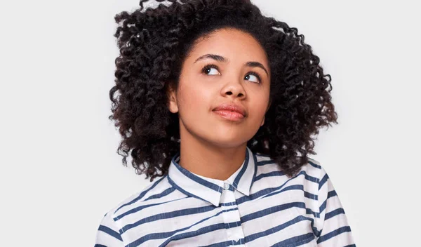 Menina muito sonhadora de pele escura vestida com camisa listrada, sentindo-se feliz, olhando para cima. Mulher afro-americana sorrindo, posando sobre a parede branca do estúdio. Conceito de emoções — Fotografia de Stock