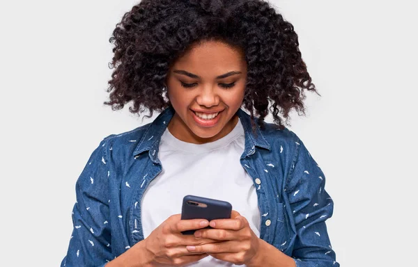 Interior close up retrato de sorrindo afro-americano jovem mulher usando celular, mensagens para sua amiga. Feliz mulher de pele escura conversando com seu namorado, olhando para a tela do telefone inteligente . — Fotografia de Stock