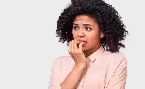 Anxious fearfull African American young woman keeps hand near mouth, feels frightened and scared, wearing in beige shirt, standing against white studio wall. Facial expressions, emotions and feelings