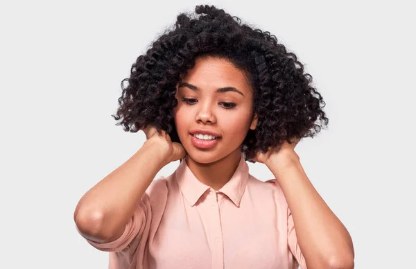 Retrato de estudio de una joven afroamericana bonita, viste una camisa beige casual, endereza su cabello afro rizado. Afro bonita hembra con una bonita sonrisa posando sobre una pared blanca. Gente emociones concepto —  Fotos de Stock
