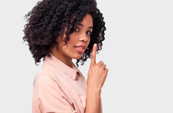 Mujer joven afroamericana confidencial vestida con camisa rosa con el dedo índice en los labios, pidiendo guardar silencio sobre la pared blanca. Hermosa hembra de piel oscura mira a la cámara pide estar tranquilo —  Fotos de Stock